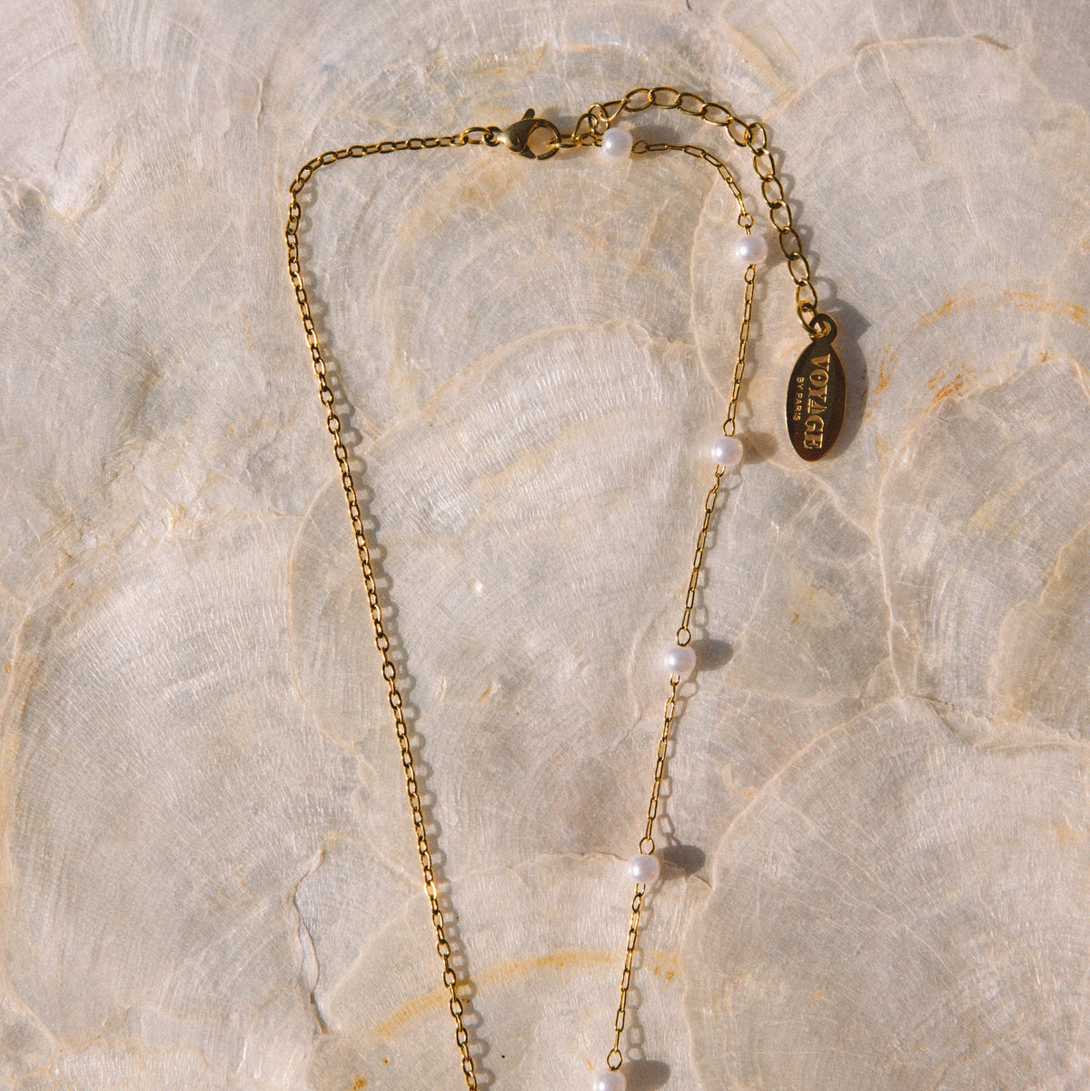 A delicate gold chain necklace with small white pearl beads and a round gold tag, laid flat on a textured beige stone surface.