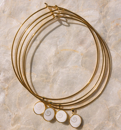 A set of four gold-plated bangle bracelets with spiral white shell charms, arranged on a textured beige stone background.