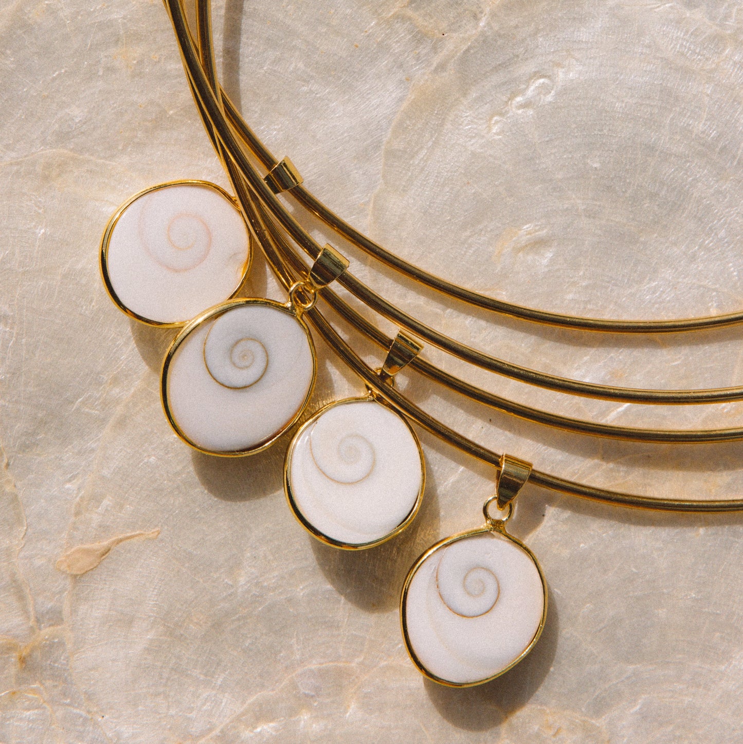 A close-up of a gold-toned multi-strand necklace with four oval white shell-like pendants featuring spiral patterns, displayed on a light beige stone surface.
