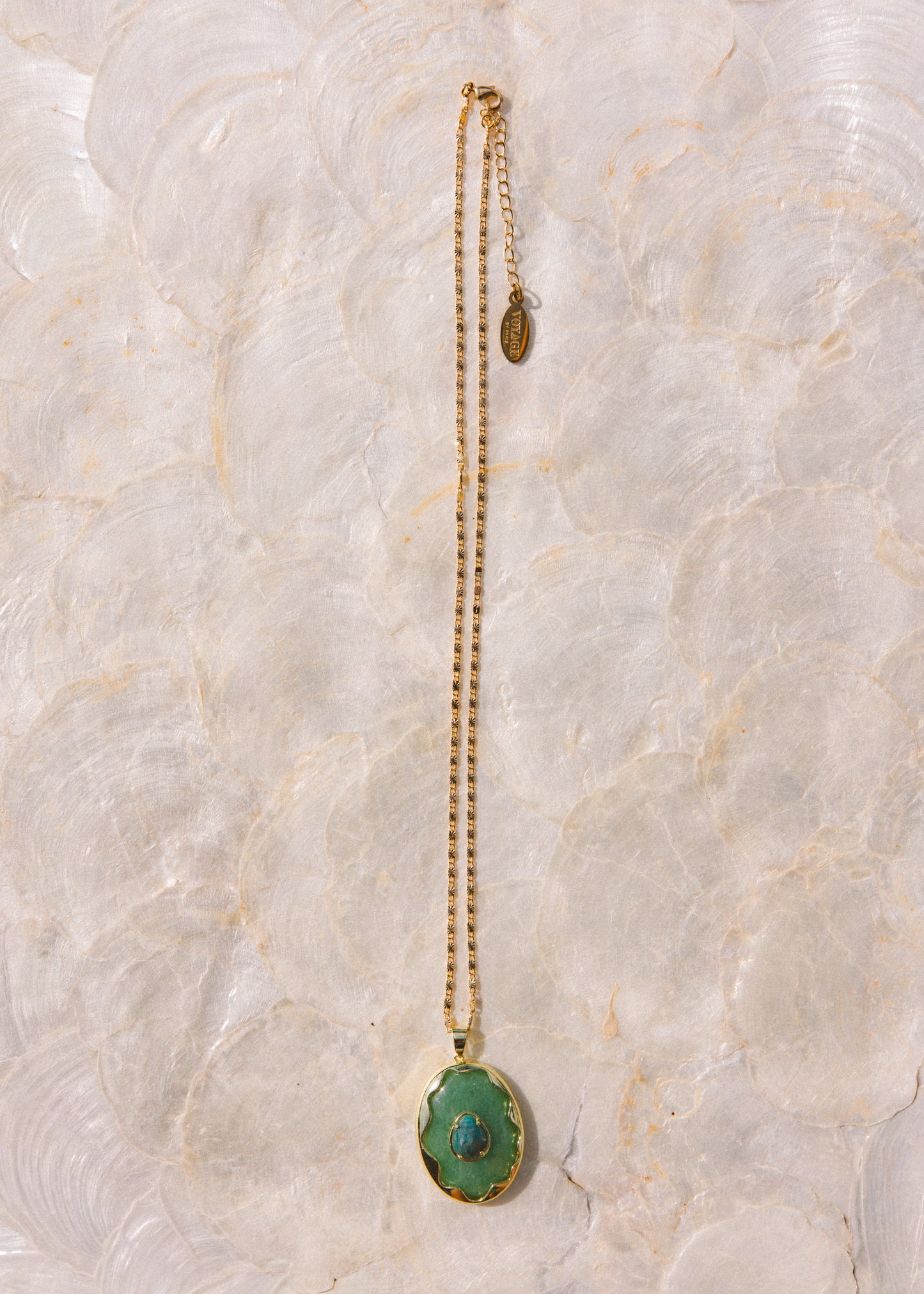 A gold chain necklace with a green oval pendant featuring a central blue stone, displayed vertically on a textured, light beige stone surface.