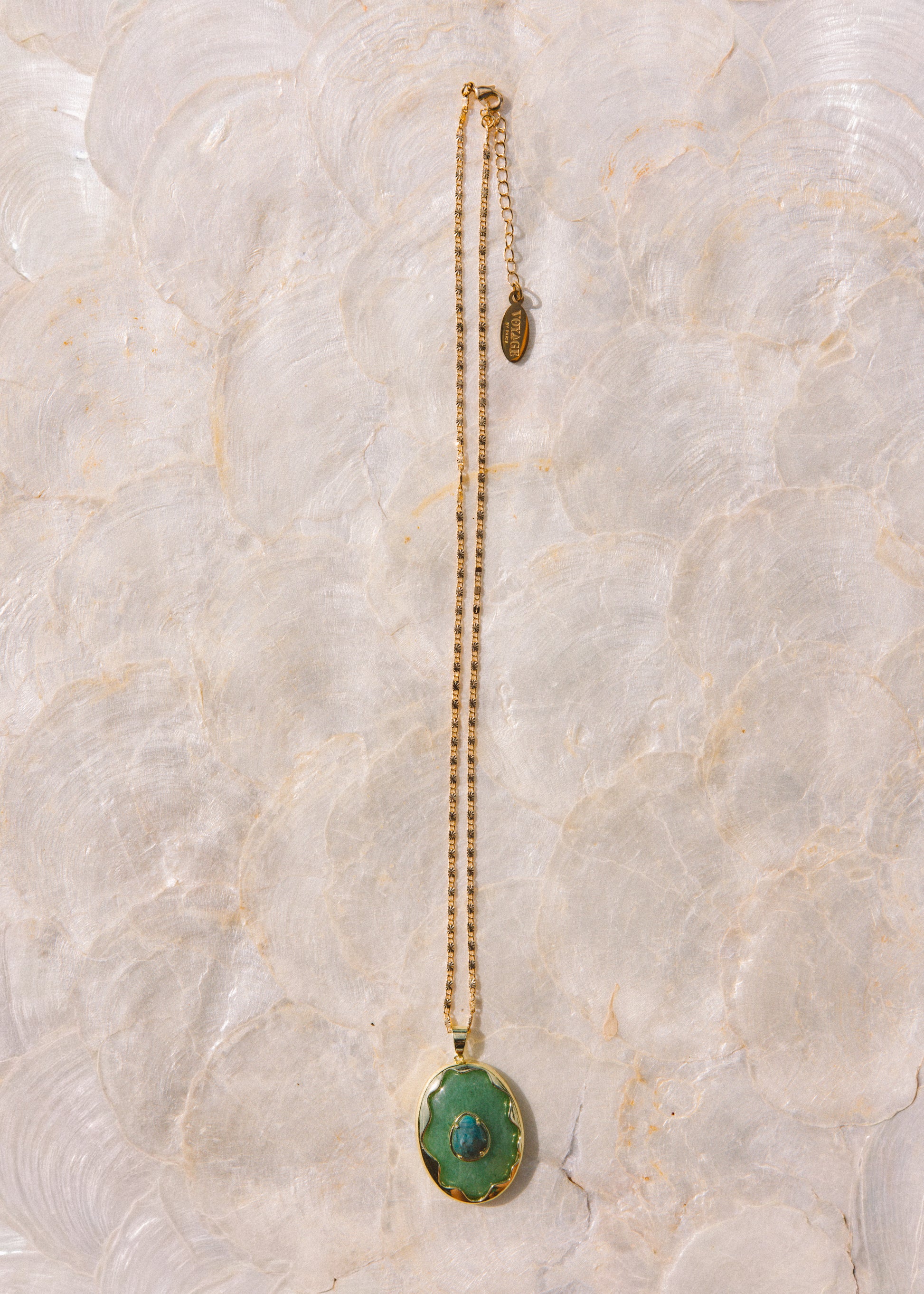 A gold chain necklace with a green oval pendant featuring a central blue stone, displayed vertically on a textured, light beige stone surface.