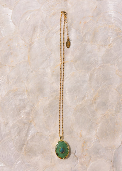 A gold chain necklace with a green oval pendant featuring a central blue stone, displayed vertically on a textured, light beige stone surface.