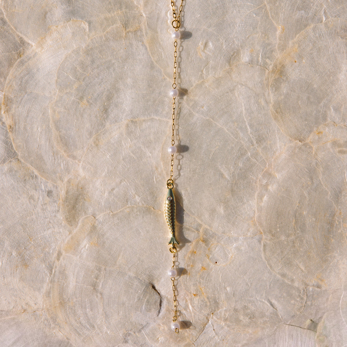 A delicate gold chain necklace featuring a small gold fish charm and clear faceted beads, laid flat on a textured beige stone surface.
