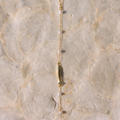 A delicate gold chain necklace featuring a small gold fish charm and clear faceted beads, laid flat on a textured beige stone surface.