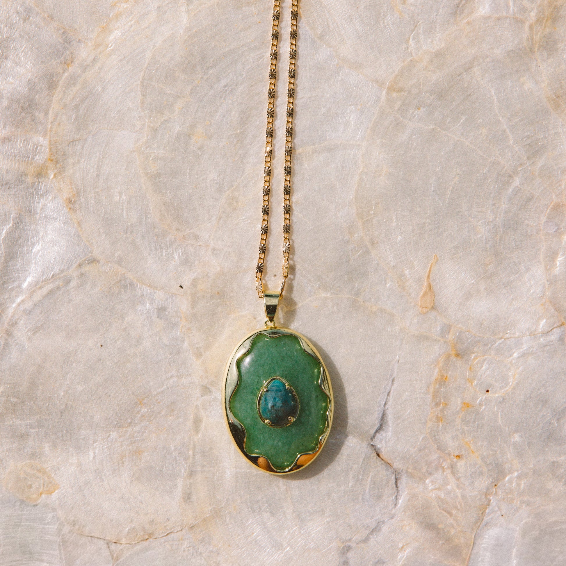 A gold chain necklace with an oval pendant featuring a green stone with a small blue teardrop inlay, displayed on a textured beige stone surface.