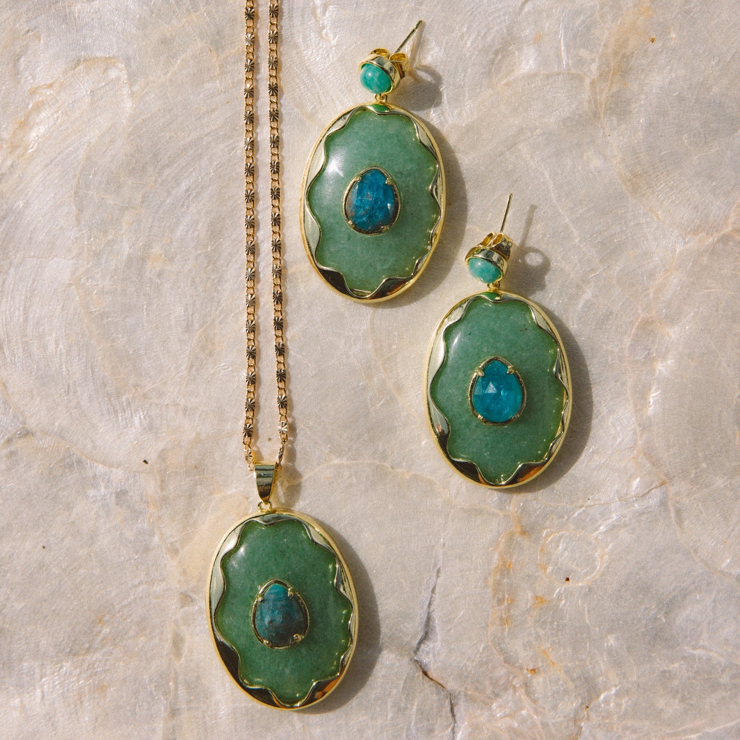 A jewelry set featuring a gold chain necklace and matching drop earrings with oval green stone pendants and small turquoise-blue center stones, arranged on a light beige stone surface.