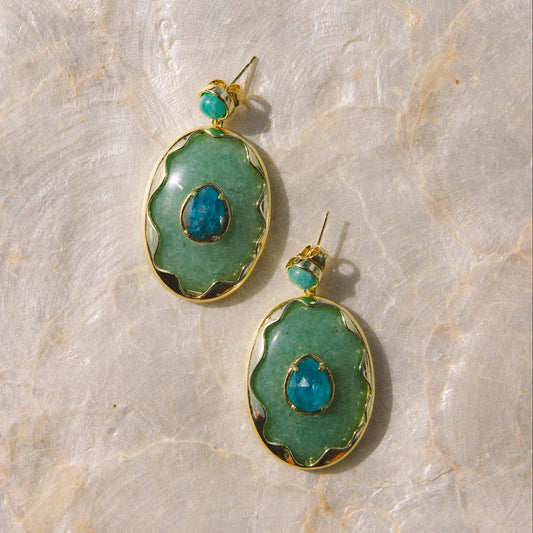 A pair of gold-tone drop earrings with scalloped green bezel settings, featuring a central faceted blue teardrop stone and a small turquoise bead above, displayed on a textured beige stone surface.