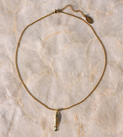 A gold-toned chain necklace featuring a small fish pendant, laid flat on a light beige marble surface.