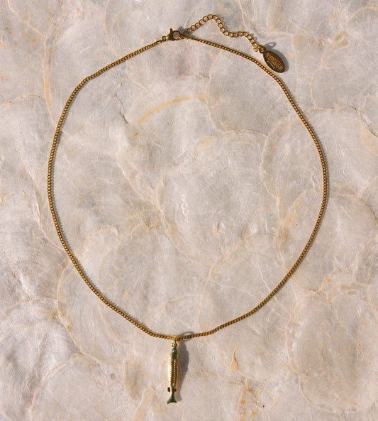 A gold-toned chain necklace featuring a small fish pendant, laid flat on a light beige marble surface.