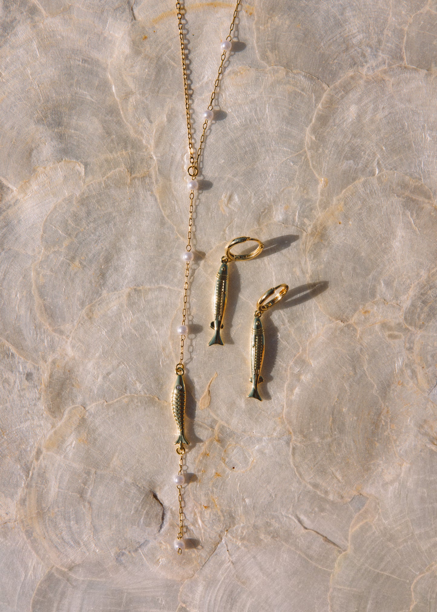 Gold-toned fish pendant necklace and matching fish-shaped earrings laid on a light beige marbled surface.