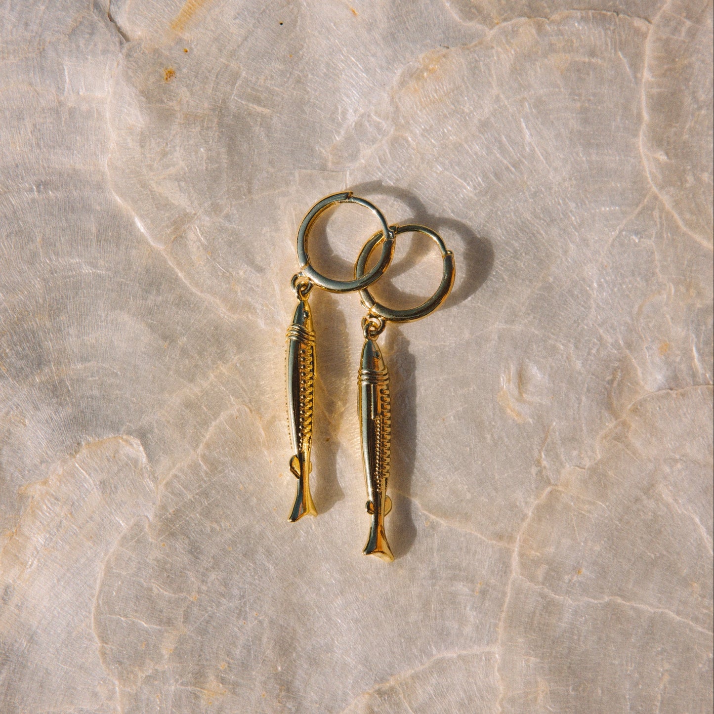 A pair of gold fish-shaped dangle earrings with matching fishhook ear wires, displayed on a light beige marble surface.