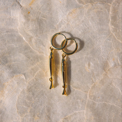 A pair of gold fish-shaped dangle earrings with matching fishhook ear wires, displayed on a light beige marble surface.