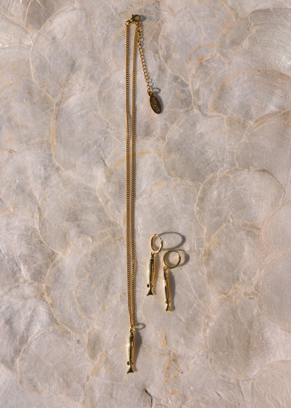 A gold-toned jewelry set featuring a long chain necklace with a small fish-shaped pendant, matching fish-shaped drop earrings, and a small ring, laid flat on a light beige marble surface.
