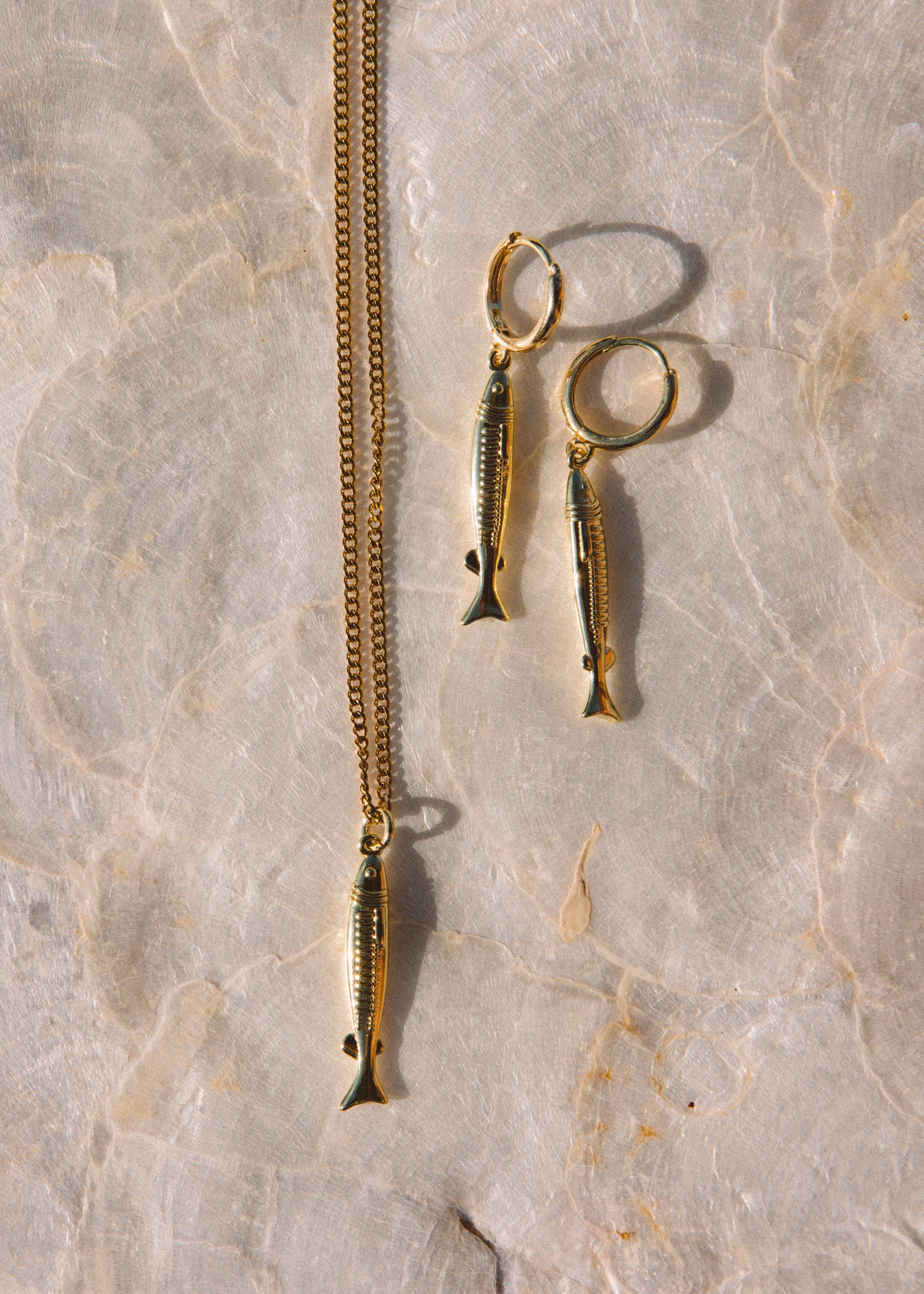 Gold-toned jewelry set featuring a chain necklace and matching hoop earrings, each with a dangling fish-shaped charm, displayed on a light beige marble surface.