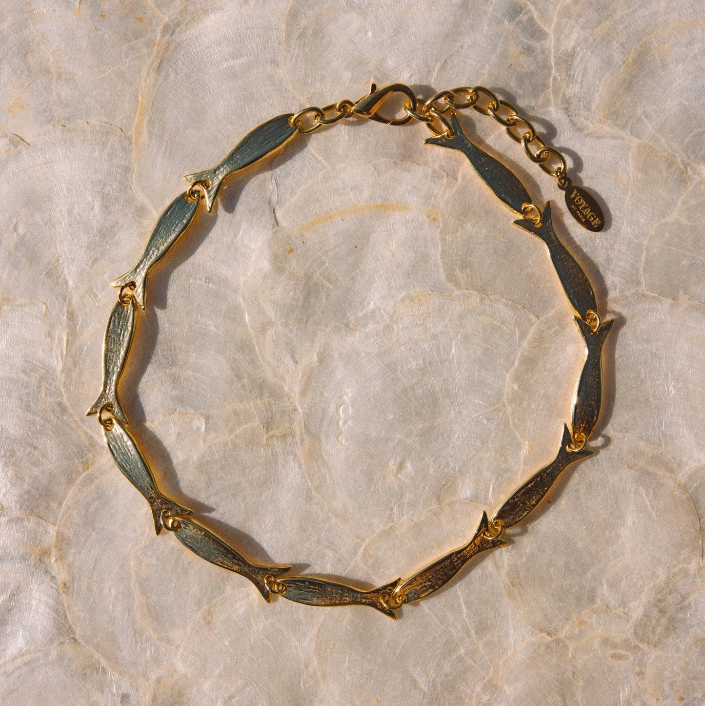 A gold-toned chain bracelet with elongated fish-shaped links, laid flat on a light beige marbled surface.