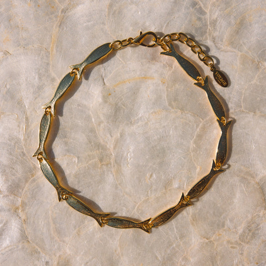 A gold-toned chain bracelet with elongated fish-shaped links, laid flat on a light beige marbled surface.