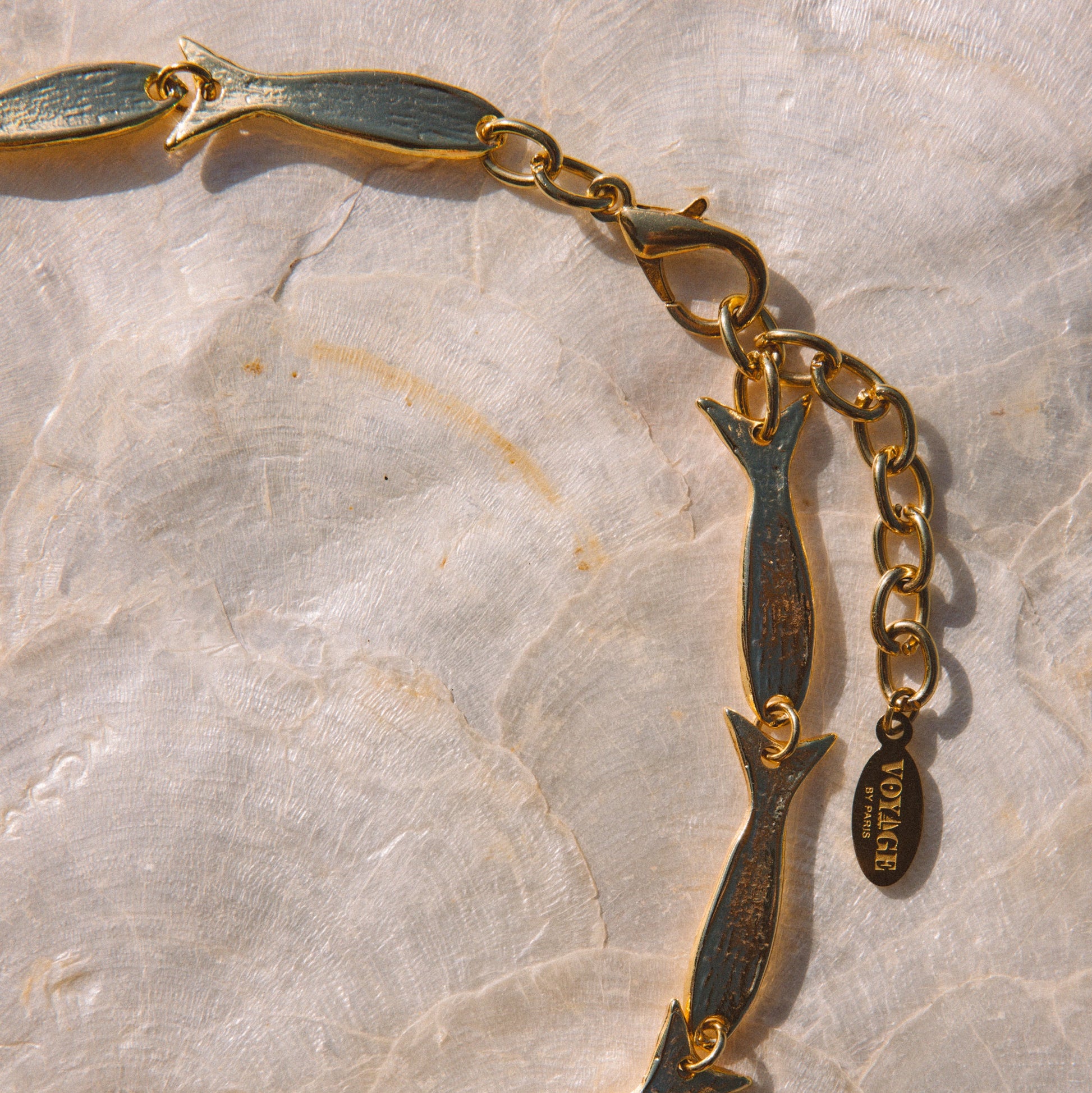 A gold-tone chain necklace featuring stylized fish charms with dark green patina detailing, displayed on a textured light beige stone surface. A small oval tag on the chain reads 'VOYAGE BY PARIS'.
