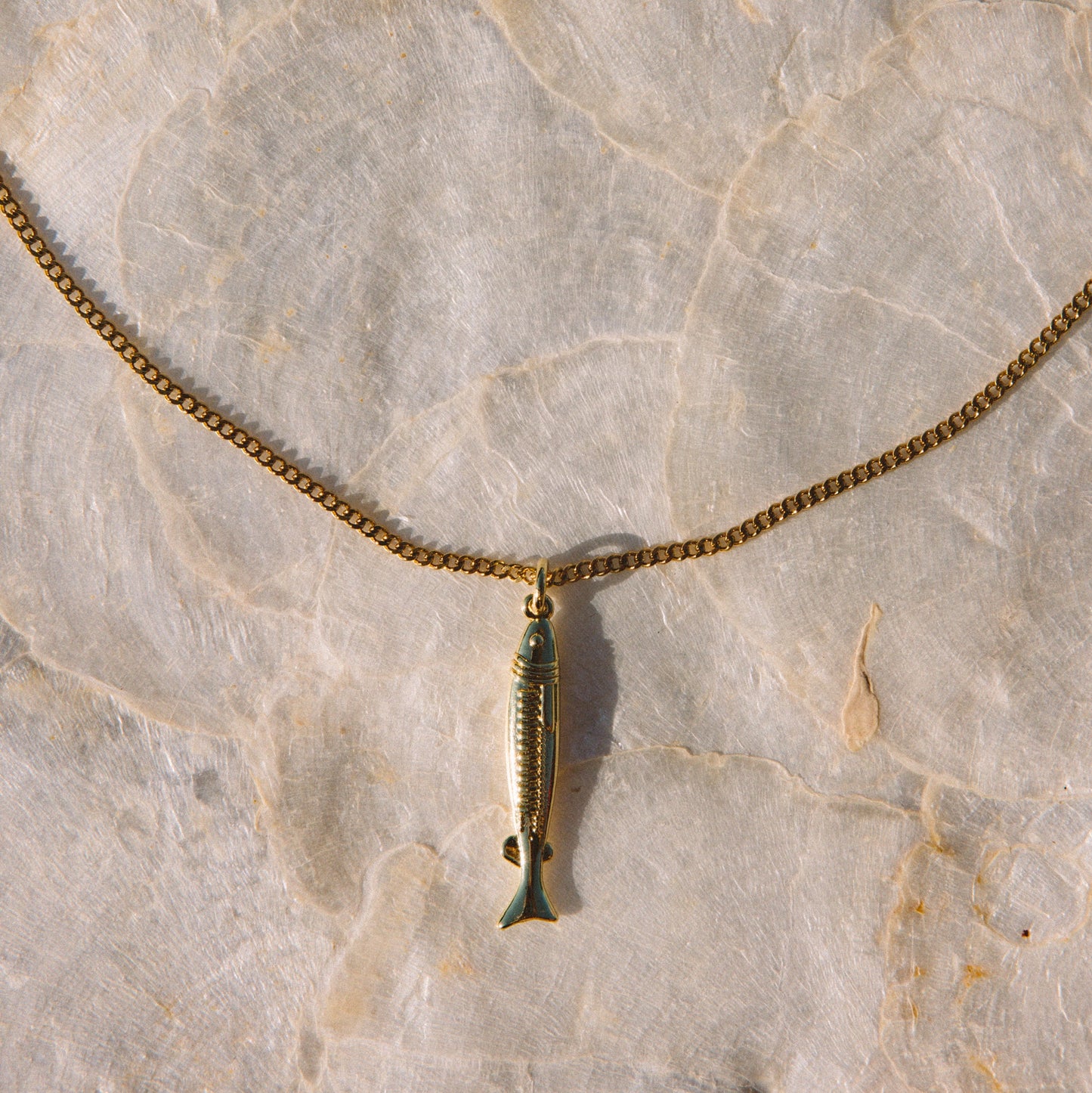 A gold-toned chain necklace with a small fish-shaped pendant, laid flat on a light beige stone surface.