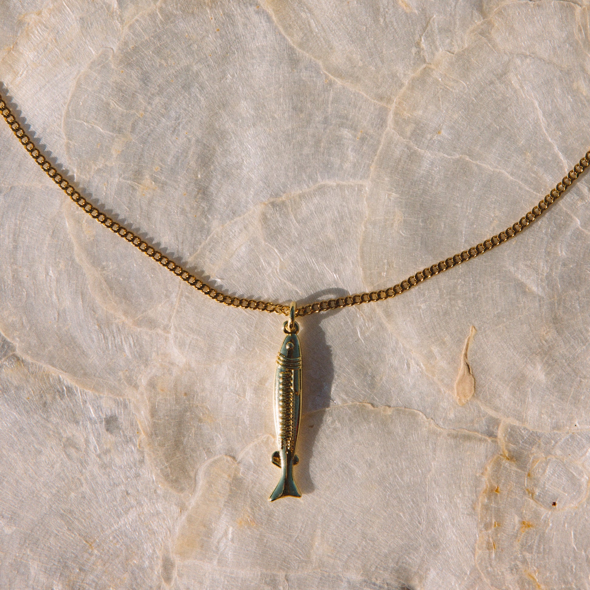 A gold-toned chain necklace with a small fish-shaped pendant, laid flat on a light beige stone surface.