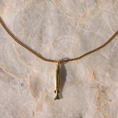 A gold-toned chain necklace with a small fish-shaped pendant, laid flat on a light beige stone surface.