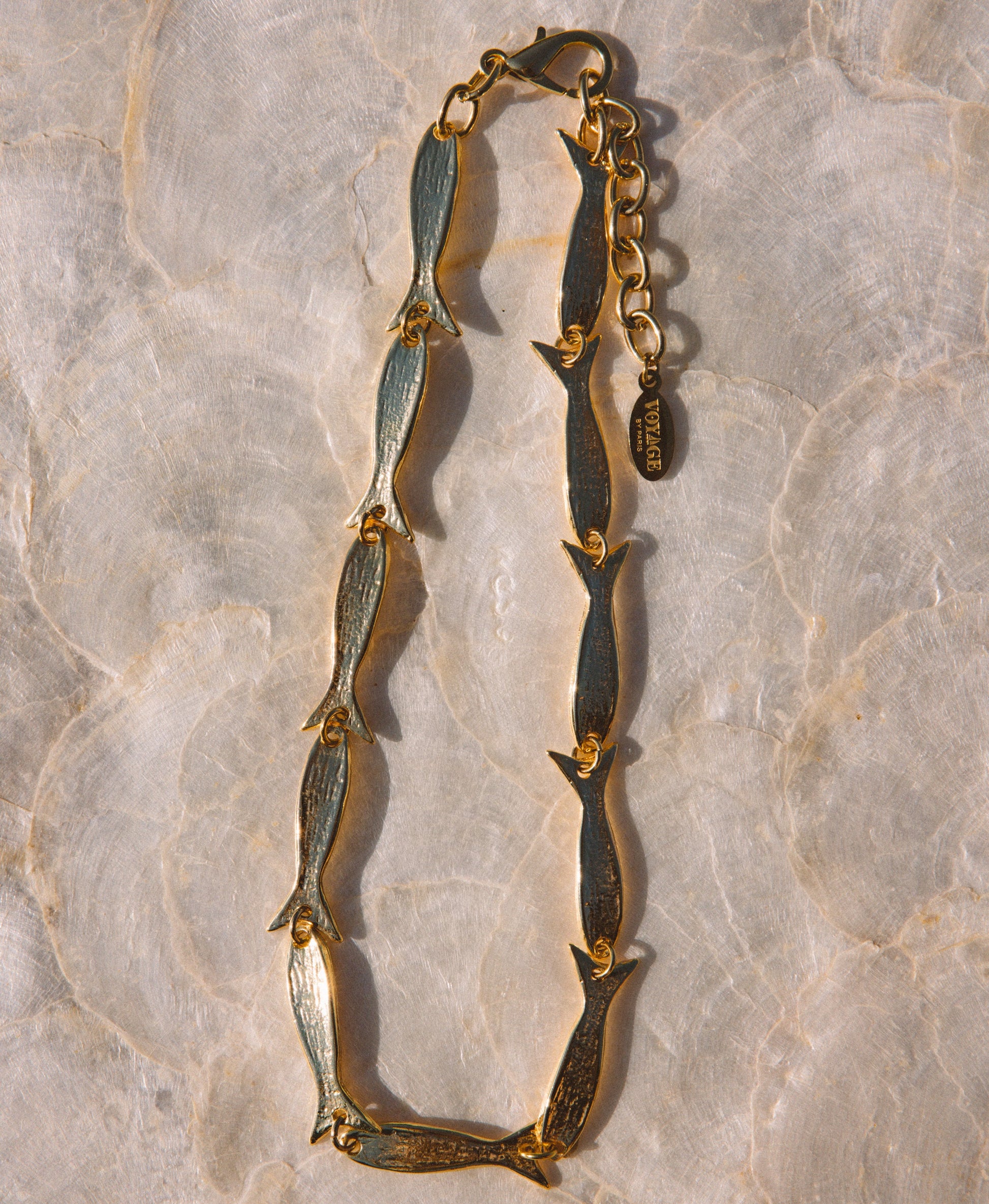 A gold-tone chain necklace featuring elongated fish-shaped links with dark green patina detailing, laid flat on a light beige marble surface.
