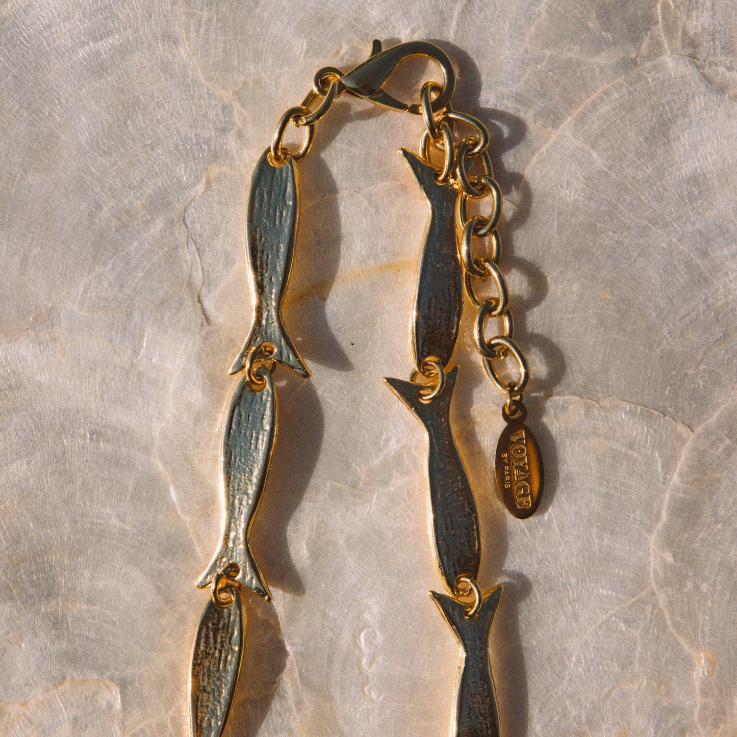 A gold-toned chain bracelet featuring elongated fish-shaped links with a textured, antiqued finish, laid flat on a light beige stone surface.