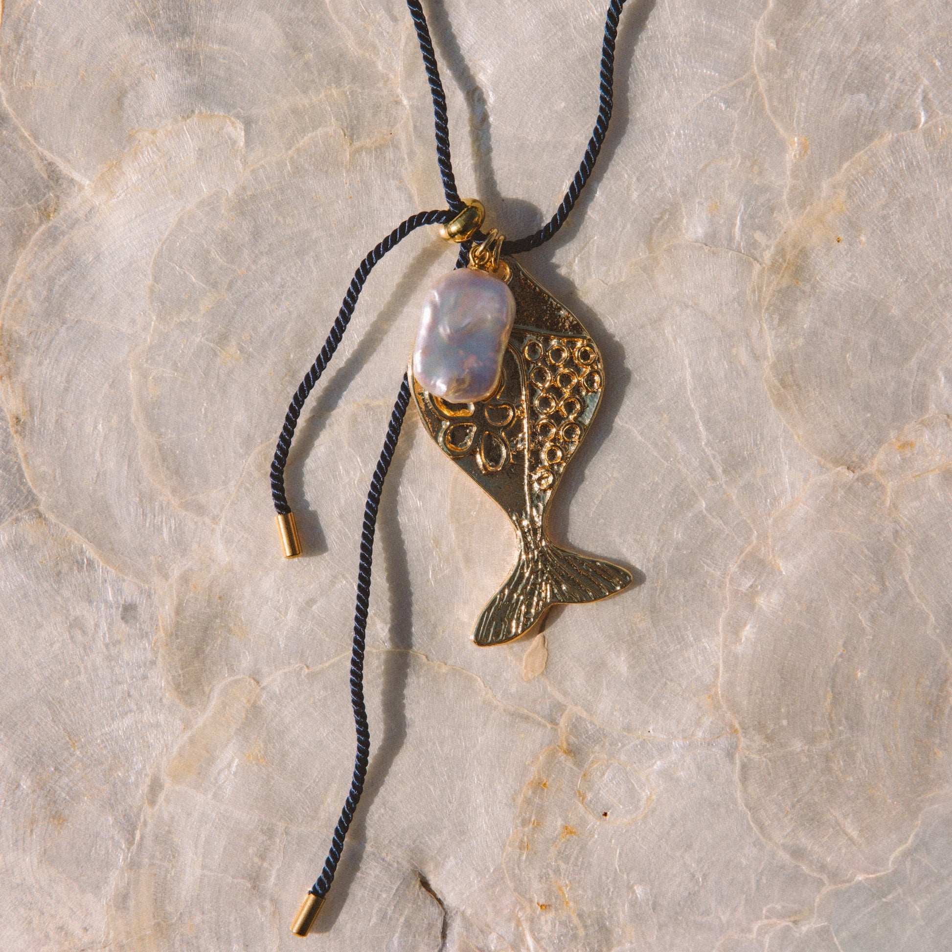 A gold-toned fish-shaped pendant necklace with a white iridescent stone, hanging from a navy blue cord on a textured beige stone surface.