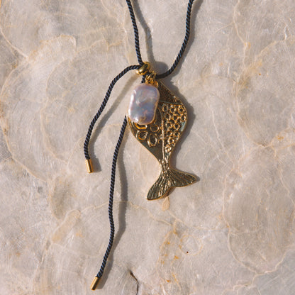 A gold-toned fish-shaped pendant necklace with a white iridescent stone, hanging from a navy blue cord on a textured beige stone surface.