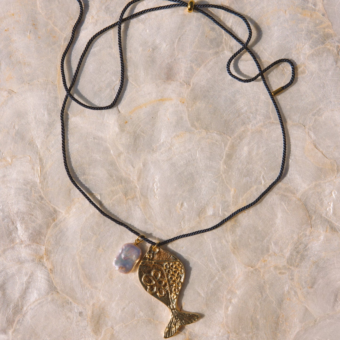 A gold-toned fish pendant necklace with a textured finish, hanging from a navy blue cord with a small iridescent bead, laid flat on a light beige stone surface.