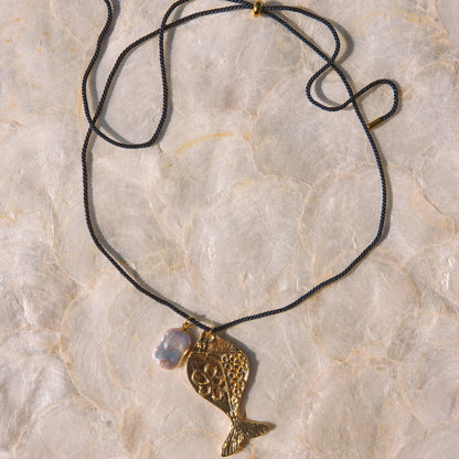 A gold-toned fish pendant necklace with a textured finish, hanging from a navy blue cord with a small iridescent bead, laid flat on a light beige stone surface.
