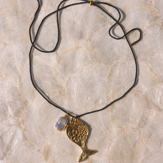 A gold-toned fish pendant necklace with a textured finish, hanging from a navy blue cord with a small iridescent bead, laid flat on a light beige stone surface.