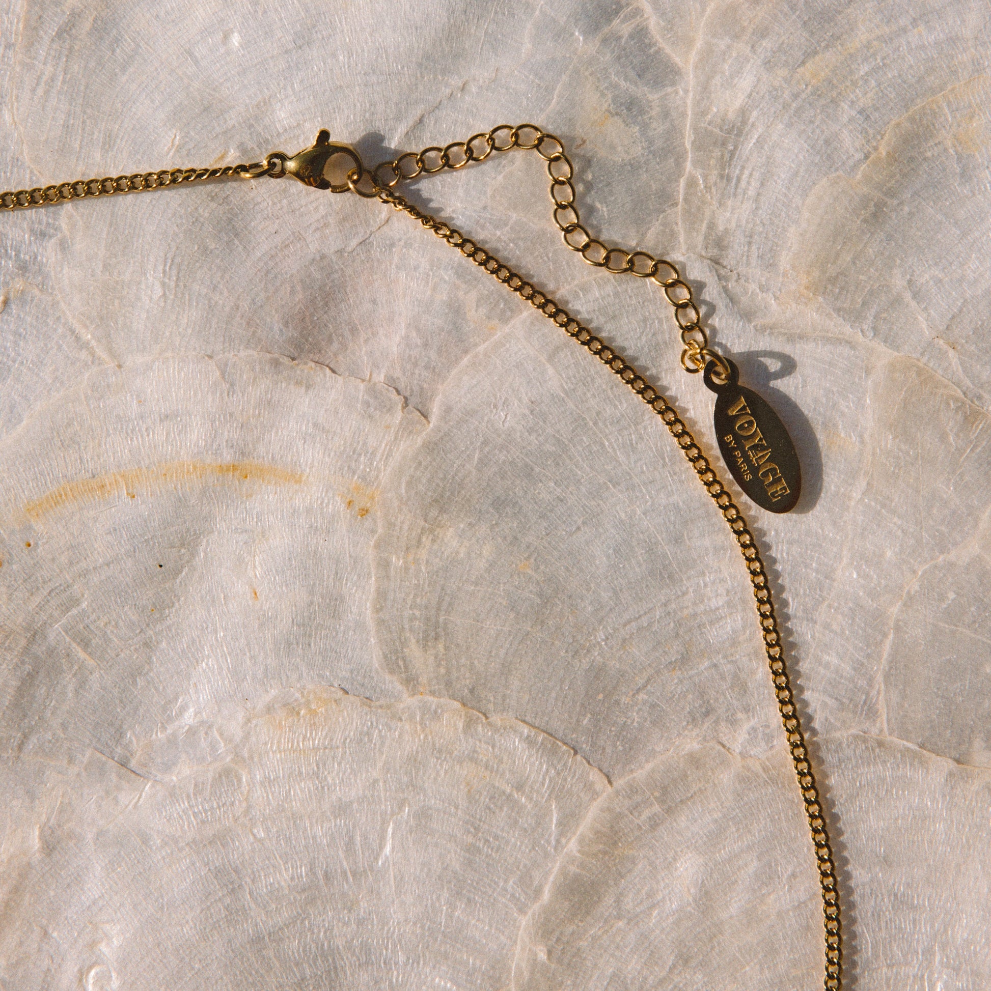 A gold chain necklace with a small oval pendant featuring the brand name 'SARAH' in gold lettering, laid out on a light beige marble surface.