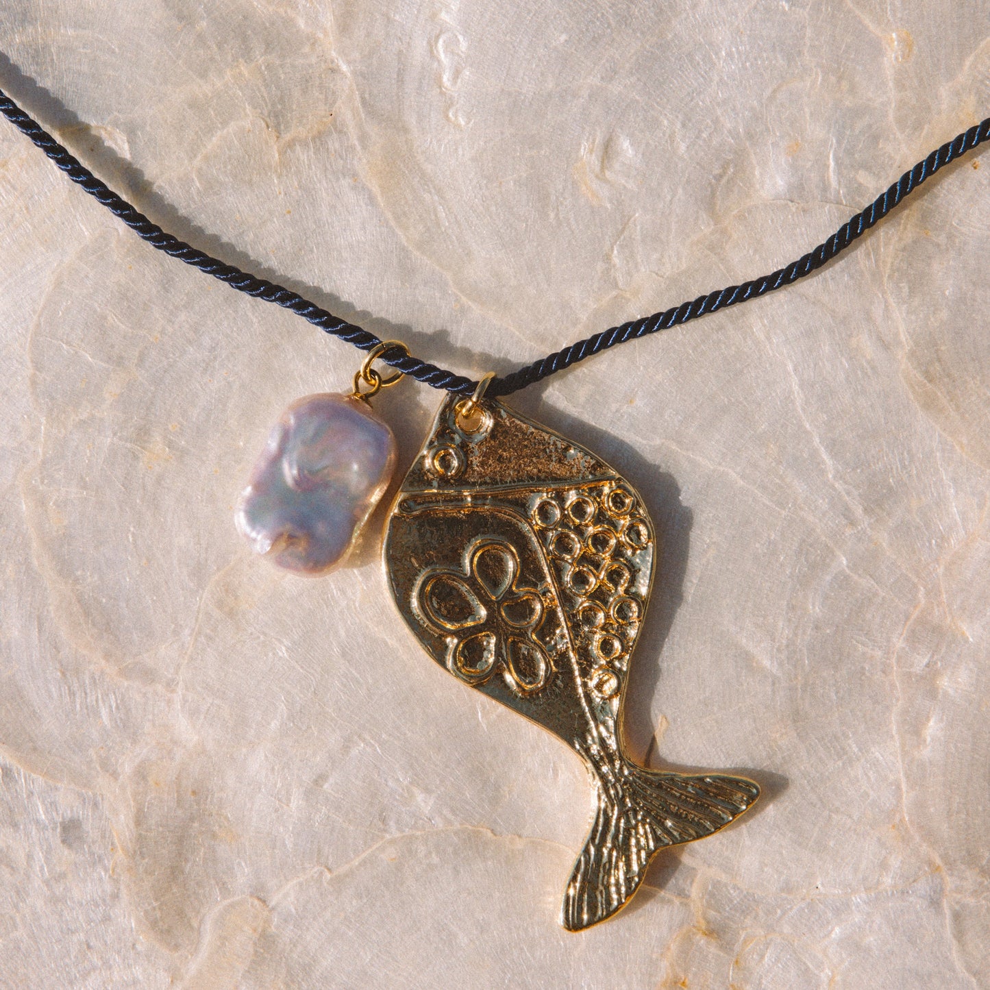 A gold-toned fish-shaped pendant necklace with a black cord and a small iridescent pearl-like bead, displayed on a light beige stone surface.