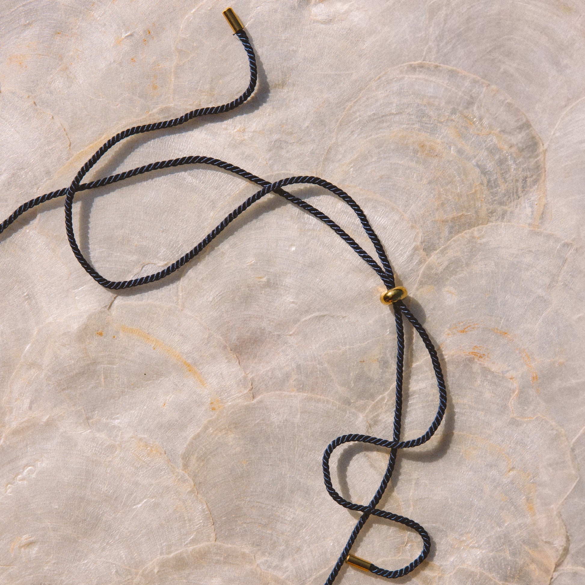 A navy blue cord necklace with small gold metal beads, laid out on a light beige marble surface.