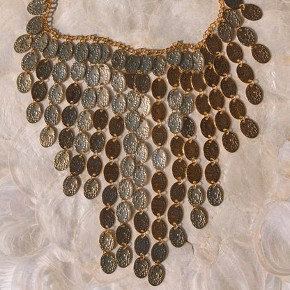 An ornate bohemian-style statement necklace featuring multiple rows of textured, coin-like oval pendants in gold and bronze tones, displayed on a soft, white textured fabric background.