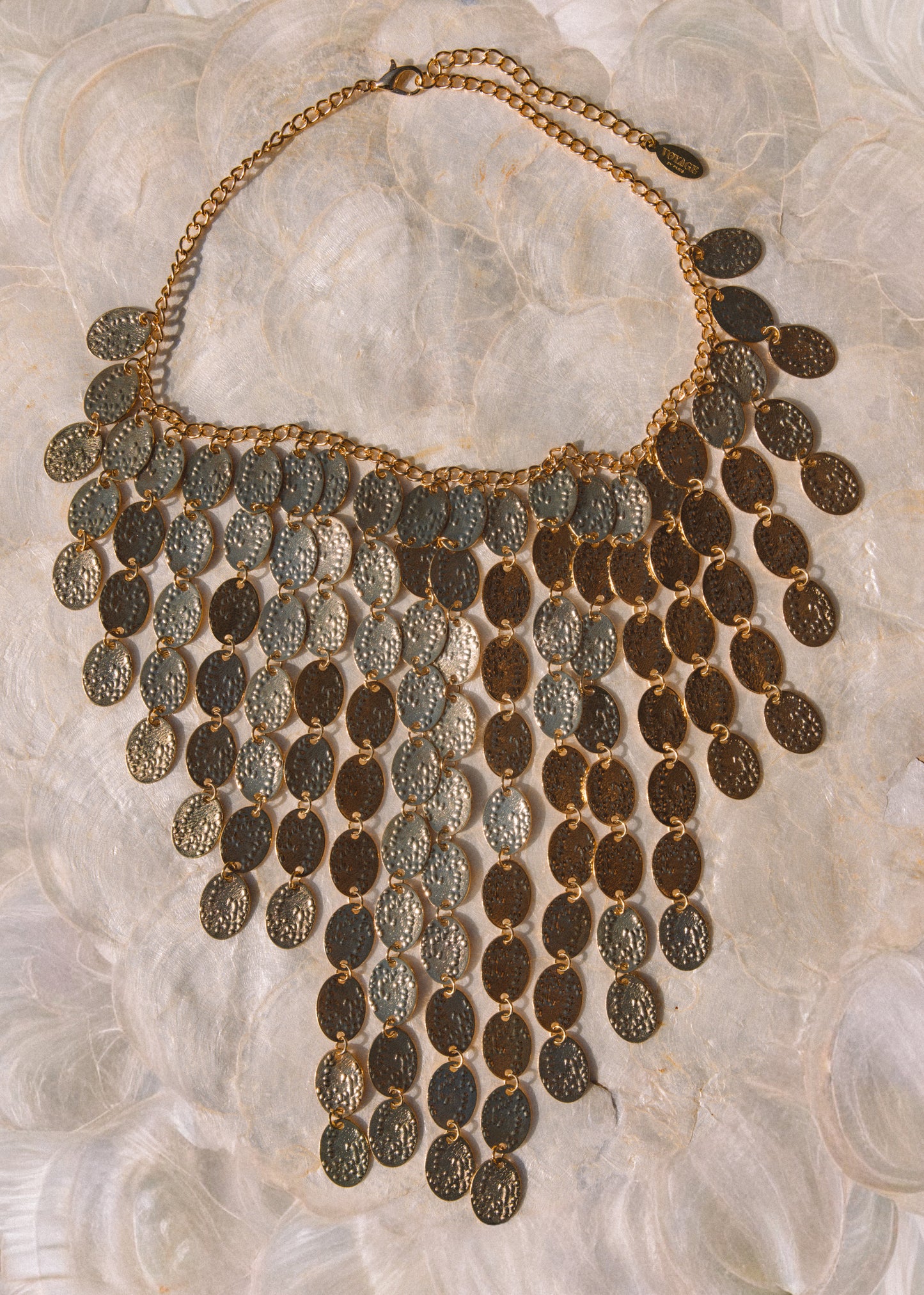 An ornate bohemian-style statement necklace featuring multiple rows of textured, coin-like oval pendants in gold and bronze tones, displayed on a soft, white textured fabric background.
