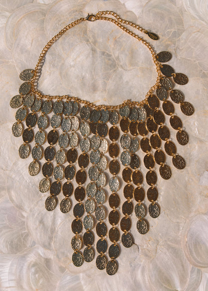 An ornate bohemian-style statement necklace featuring multiple rows of textured, coin-like oval pendants in gold and bronze tones, displayed on a soft, white textured fabric background.