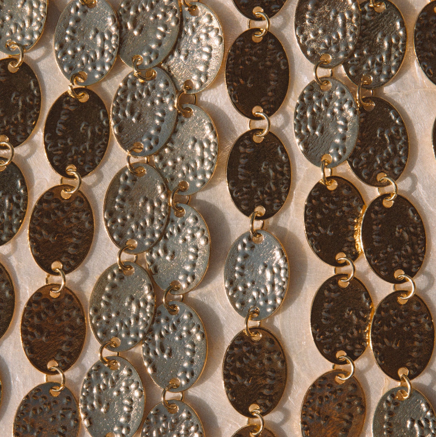 Close-up of multiple strands of oval link jewelry with hammered metallic finishes in silver, bronze, and dark brown tones, arranged vertically against a light neutral background.