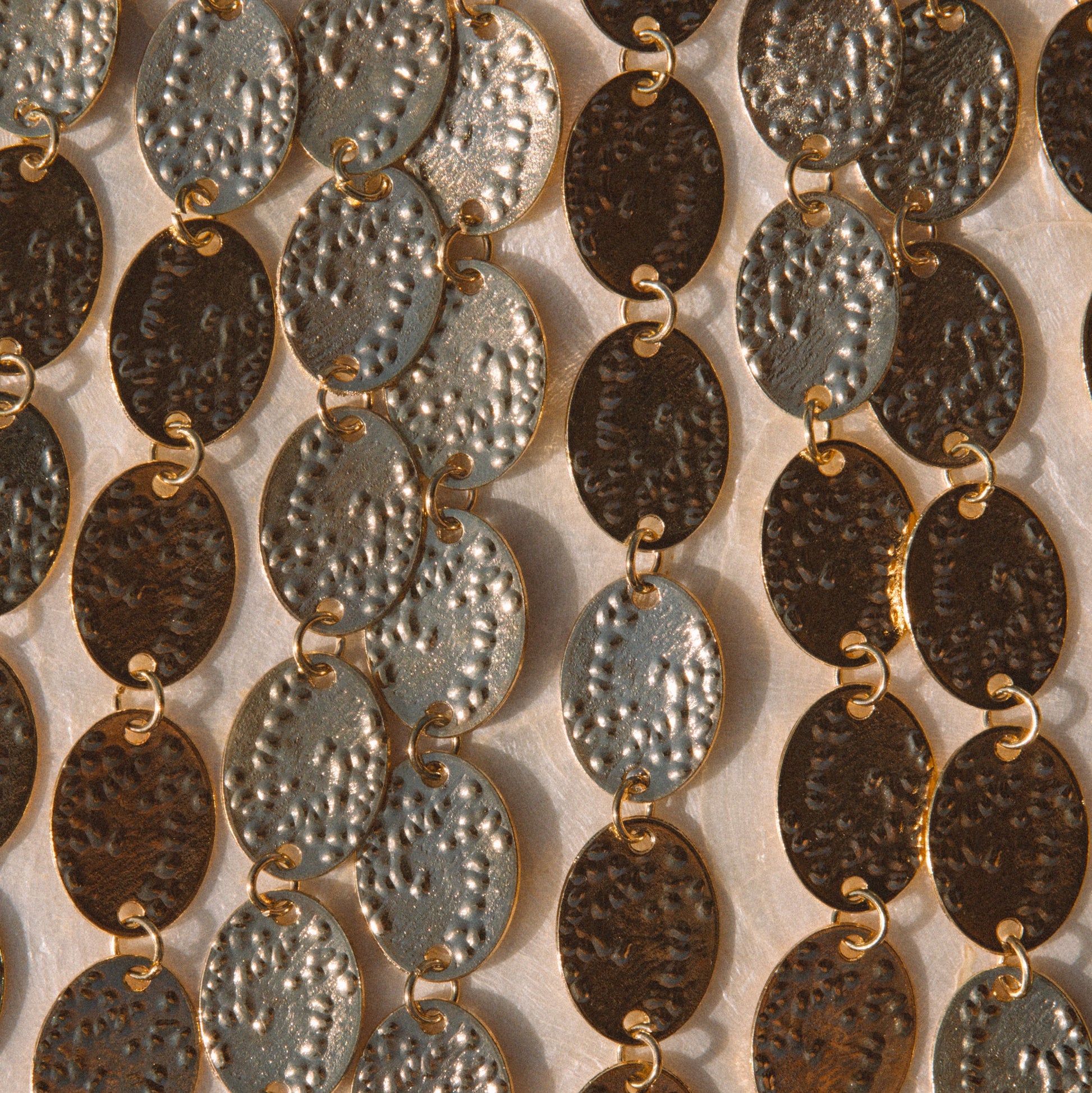 Close-up of multiple strands of oval link jewelry with hammered metallic finishes in silver, bronze, and dark brown tones, arranged vertically against a light neutral background.