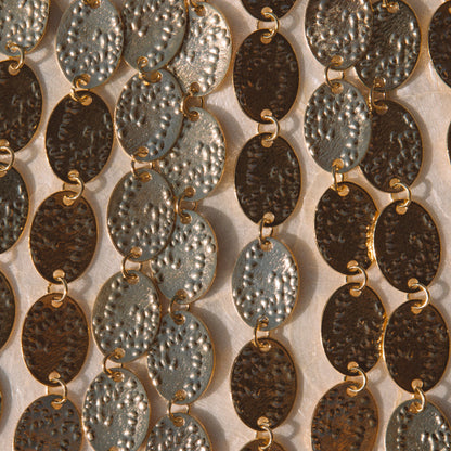 Close-up of multiple strands of oval link jewelry with hammered metallic finishes in silver, bronze, and dark brown tones, arranged vertically against a light neutral background.