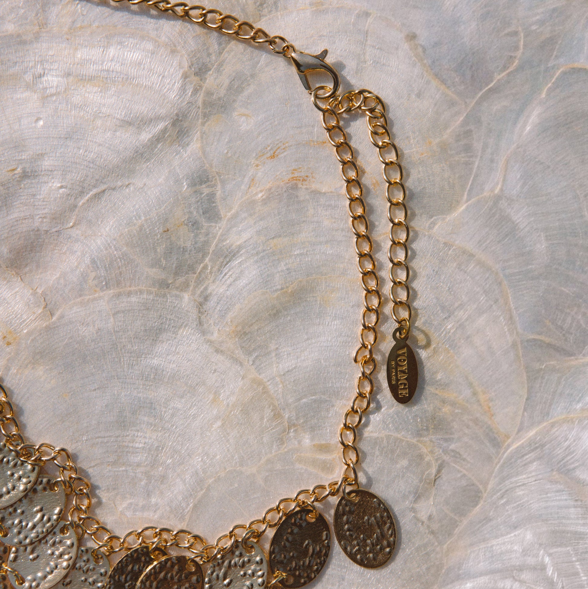 A gold-toned chain necklace with multiple overlapping textured disc charms, laid flat on a light marbled surface. A small hanging tag is visible near the clasp.