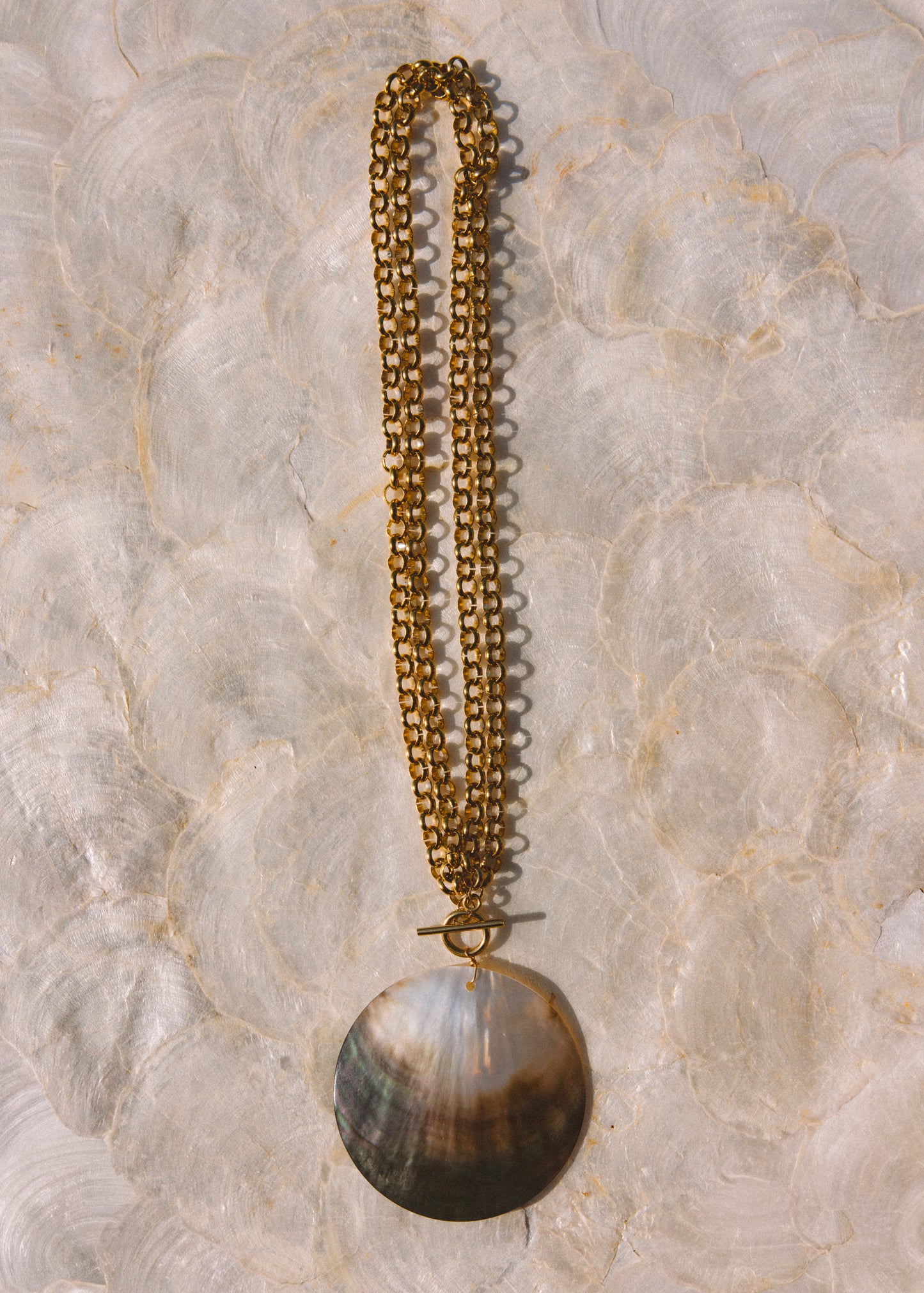 A double-strand gold chain necklace with a large circle pendant, laid flat on a textured, light-colored stone surface.