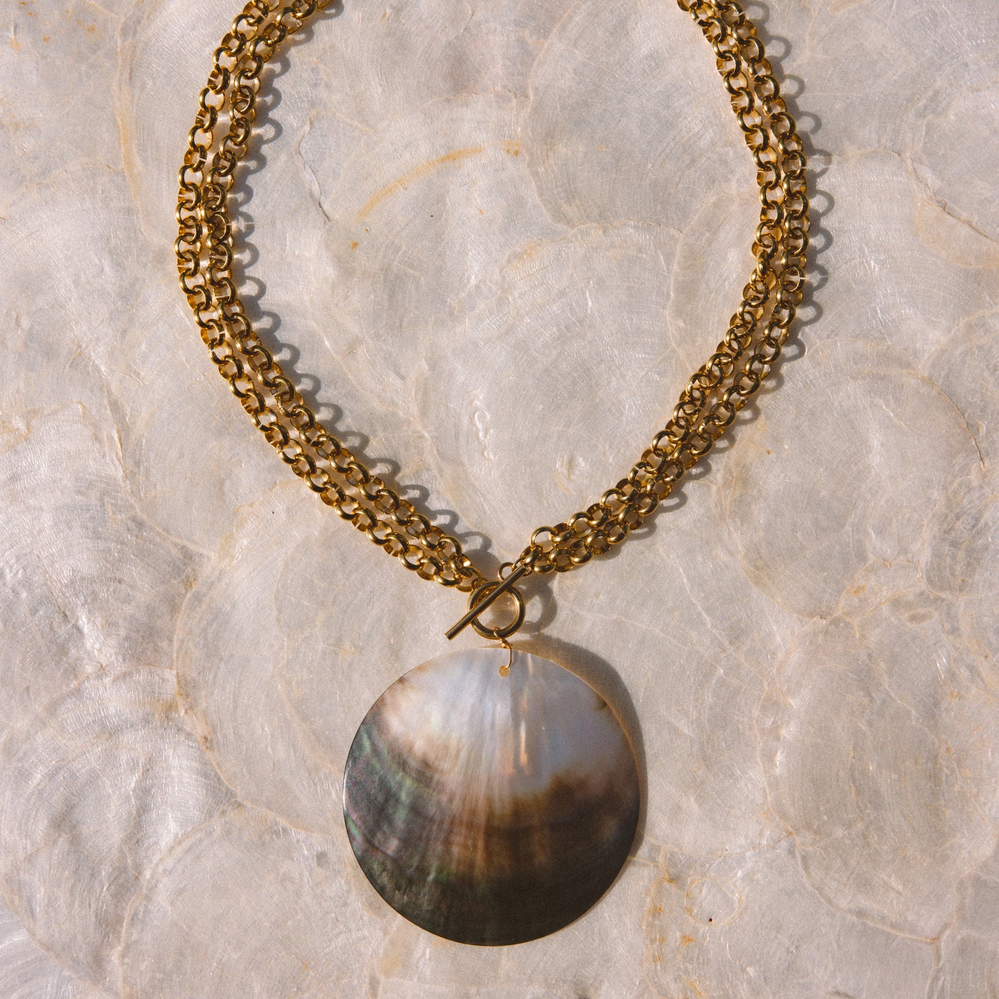 A gold-tone chain necklace with a large, iridescent round pendant, laid flat on a textured beige stone surface.