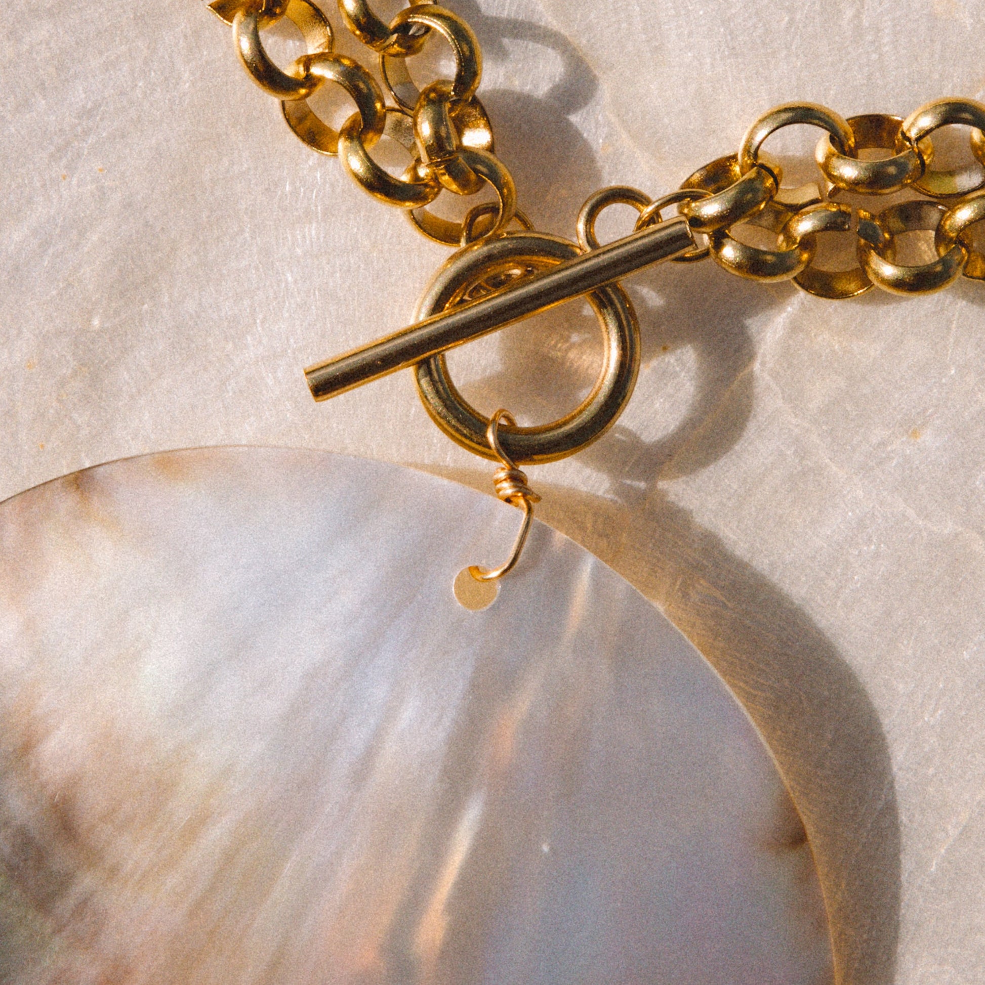 Close-up of a gold chain necklace with a toggle clasp and a large circle pendant in iridescent white, beige, and brown tones, resting on a textured light surface.