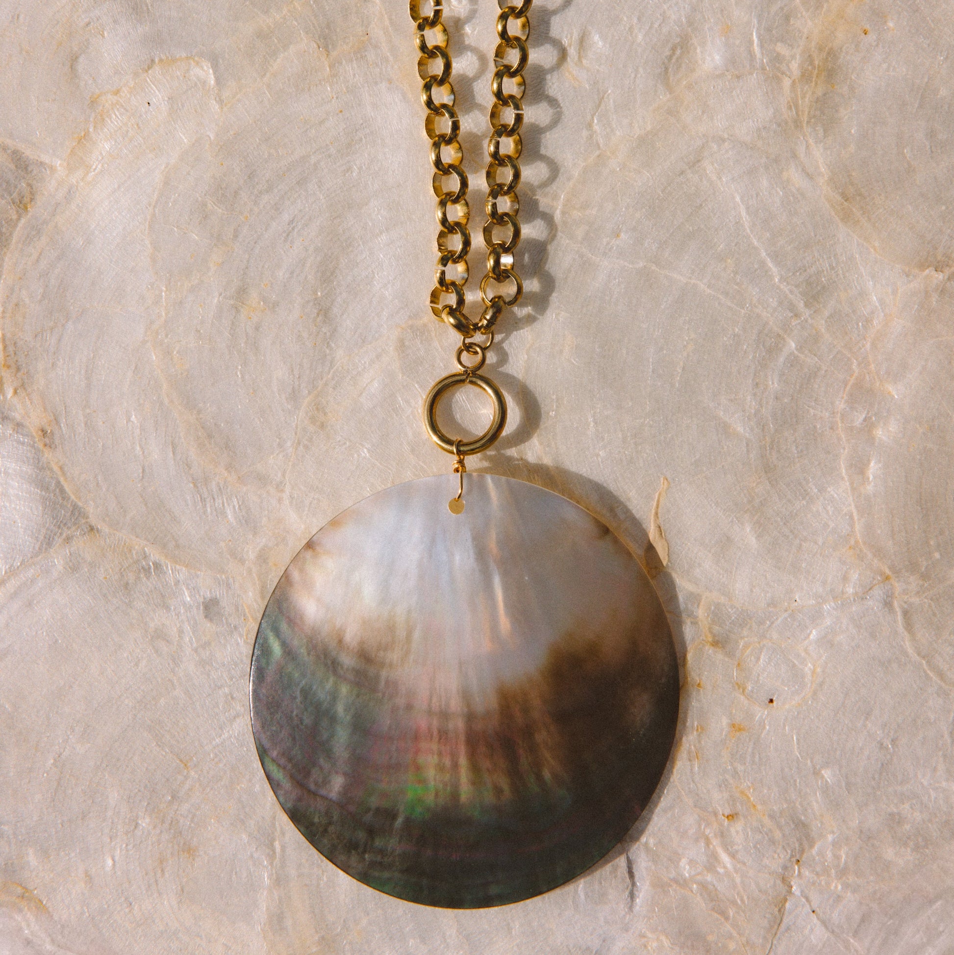 A gold-tone chain necklace with a large pendant featuring iridescent brown, green, and cream tones, displayed on a textured beige stone surface.