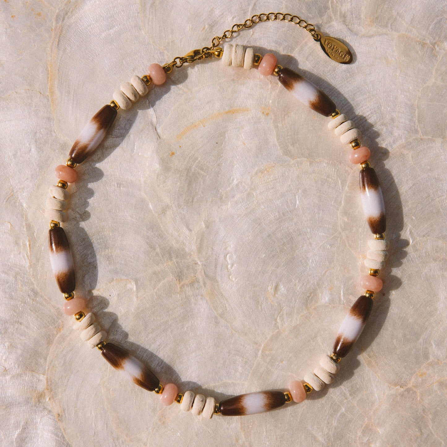 A bohemian-style beaded necklace with alternating white beads, brown-and-white marbled beads, and small pink beads, finished with a gold-tone clasp and extender chain, displayed on a textured beige surface.