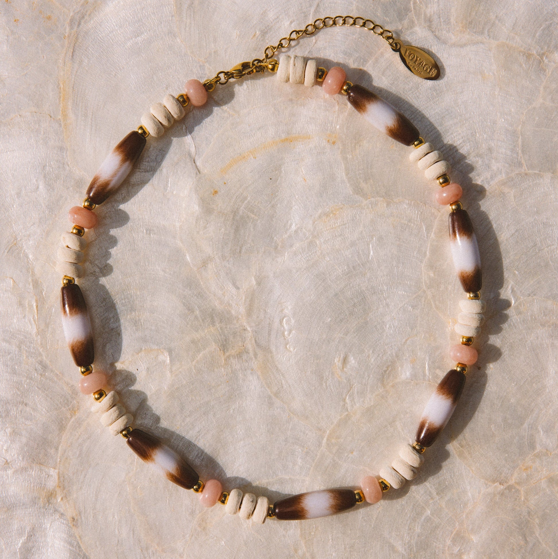 A bohemian-style beaded necklace with alternating white beads, brown-and-white marbled beads, and small pink beads, finished with a gold-tone clasp and extender chain, displayed on a textured beige surface.