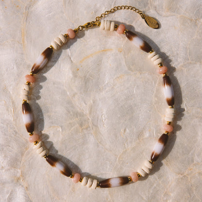 A bohemian-style beaded necklace with alternating white beads, brown-and-white marbled beads, and small pink beads, finished with a gold-tone clasp and extender chain, displayed on a textured beige surface.