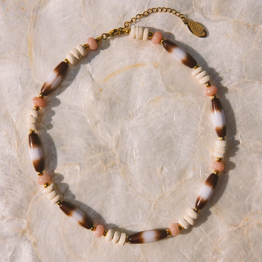 A bohemian-style beaded necklace with alternating white beads, brown-and-white marbled beads, and small pink beads, finished with a gold-tone clasp and extender chain, displayed on a textured beige surface.