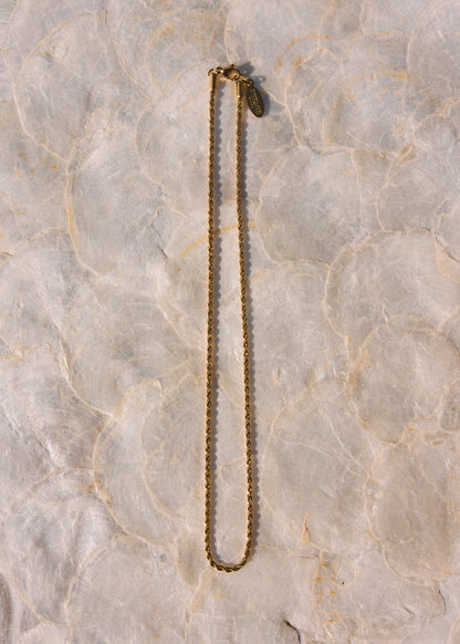 A gold-toned chain necklace with a small dark tag near the clasp, laid flat on a light beige marbled surface.